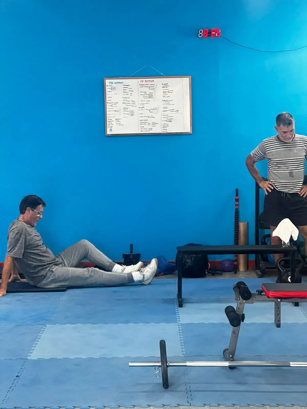 Un profe acompañando a dos personas durante el entrenamiento, frente a la pizarra de rutinas